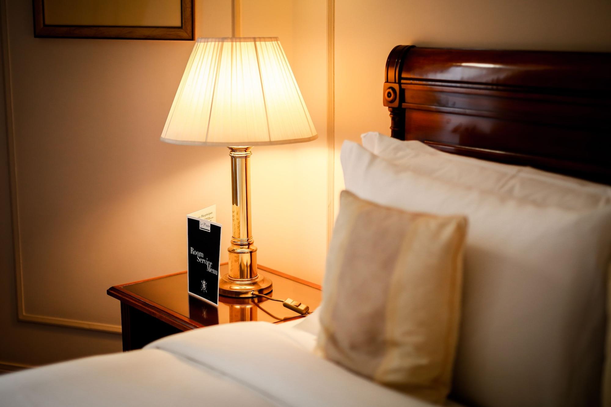 布鲁塞尔广场酒店  外观 照片 The photo shows a cozy bedside scene in a well-decorated bedroom. There is a wooden bed with neatly arranged white pillows and a soft, beige decorative cushion. Beside the bed, there's a polished wooden nightstand with a warm, glowing lamp featuring 
