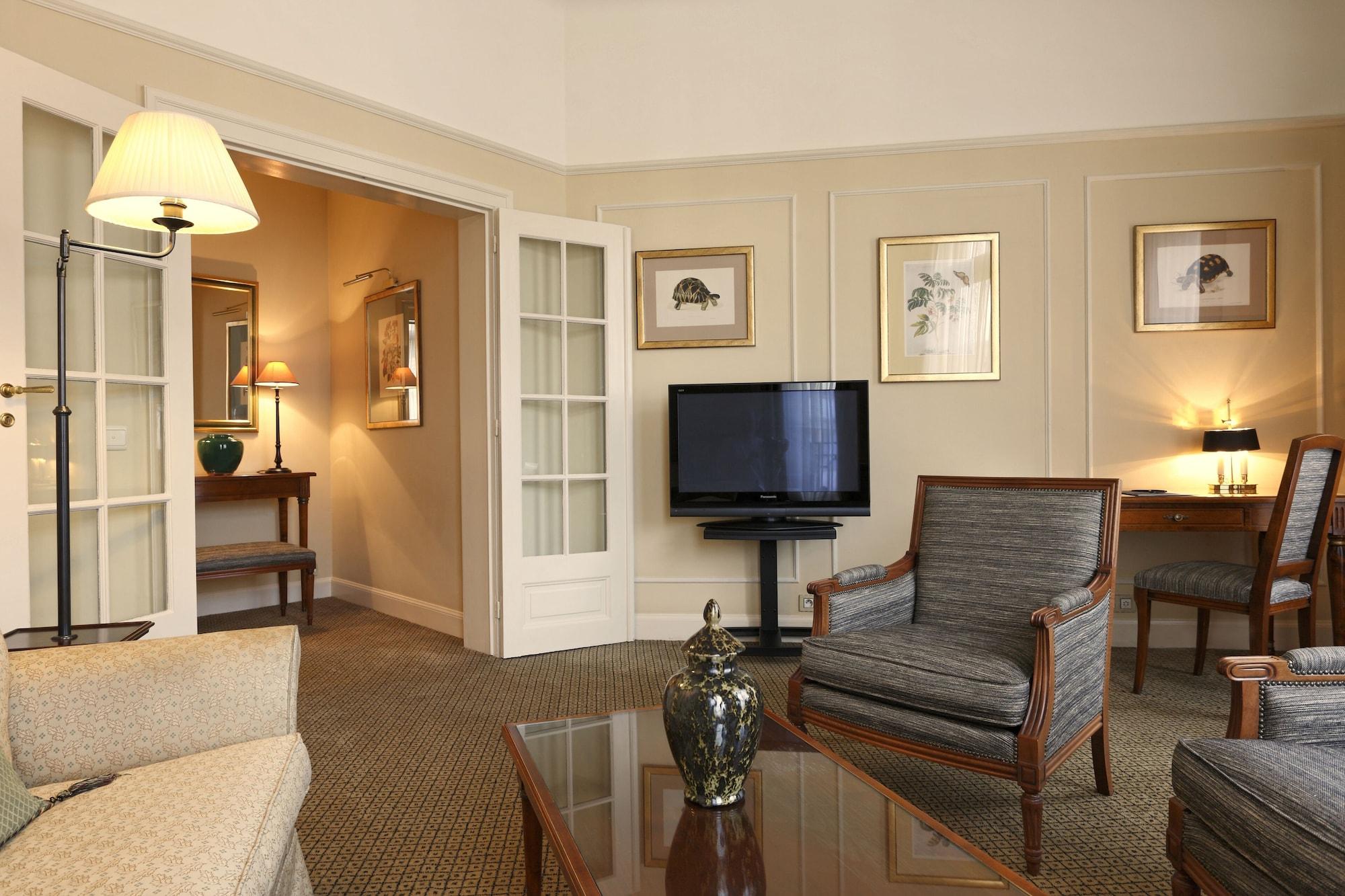 布鲁塞尔广场酒店  客房 照片 The image shows a cozy and elegantly designed living room area. There is a comfortable sofa and armchairs arranged around a coffee table. A large television is mounted on a stand, facing the seating area. The walls are adorned with framed artwork, an