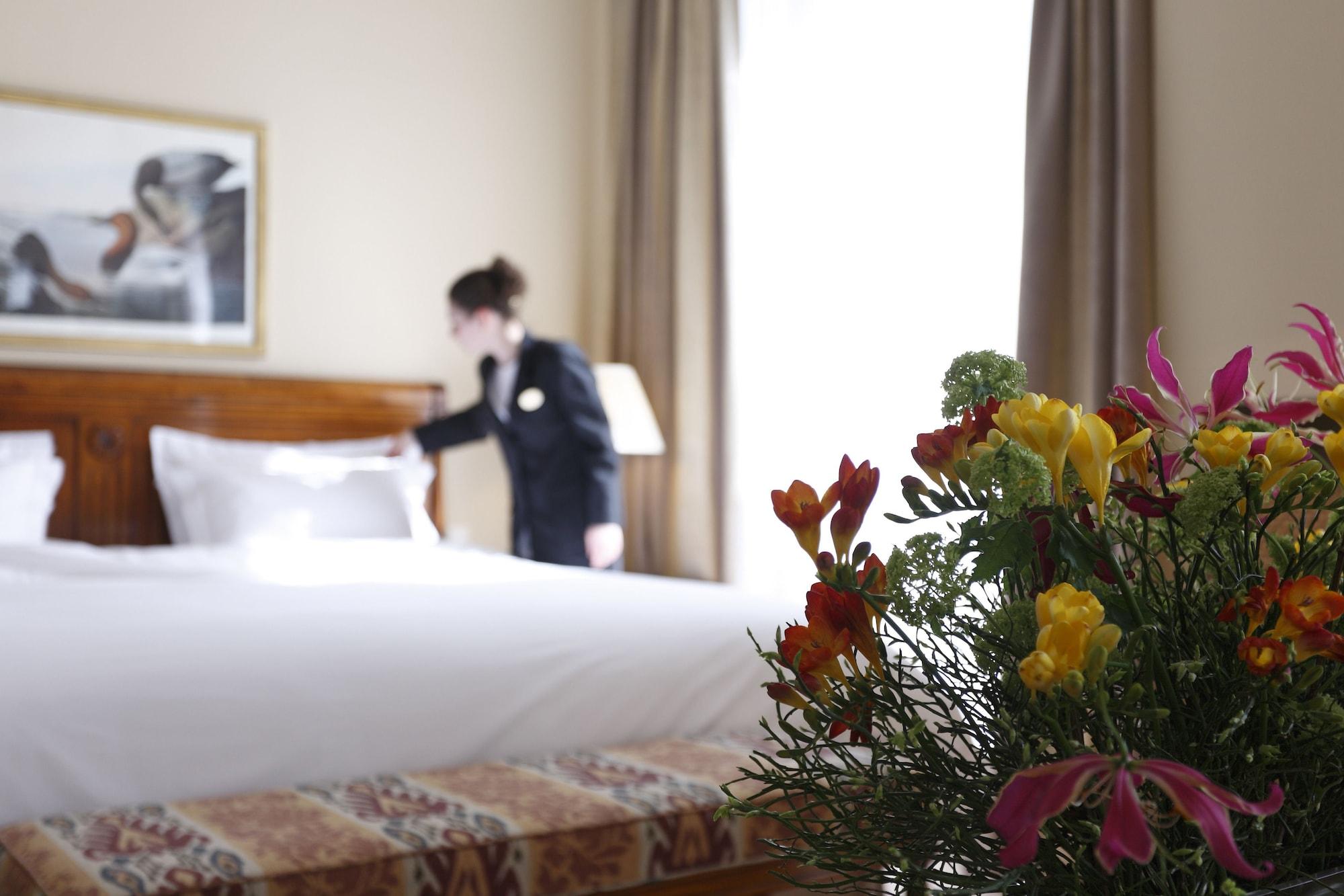 布鲁塞尔广场酒店  外观 照片 The photo shows a hotel room with a neatly made bed in the background. A staff member, dressed in a uniform, is fixing the bed. In the foreground, there is a bouquet of colorful flowers, adding a touch of vibrancy to the scene. The room is well-lit, 