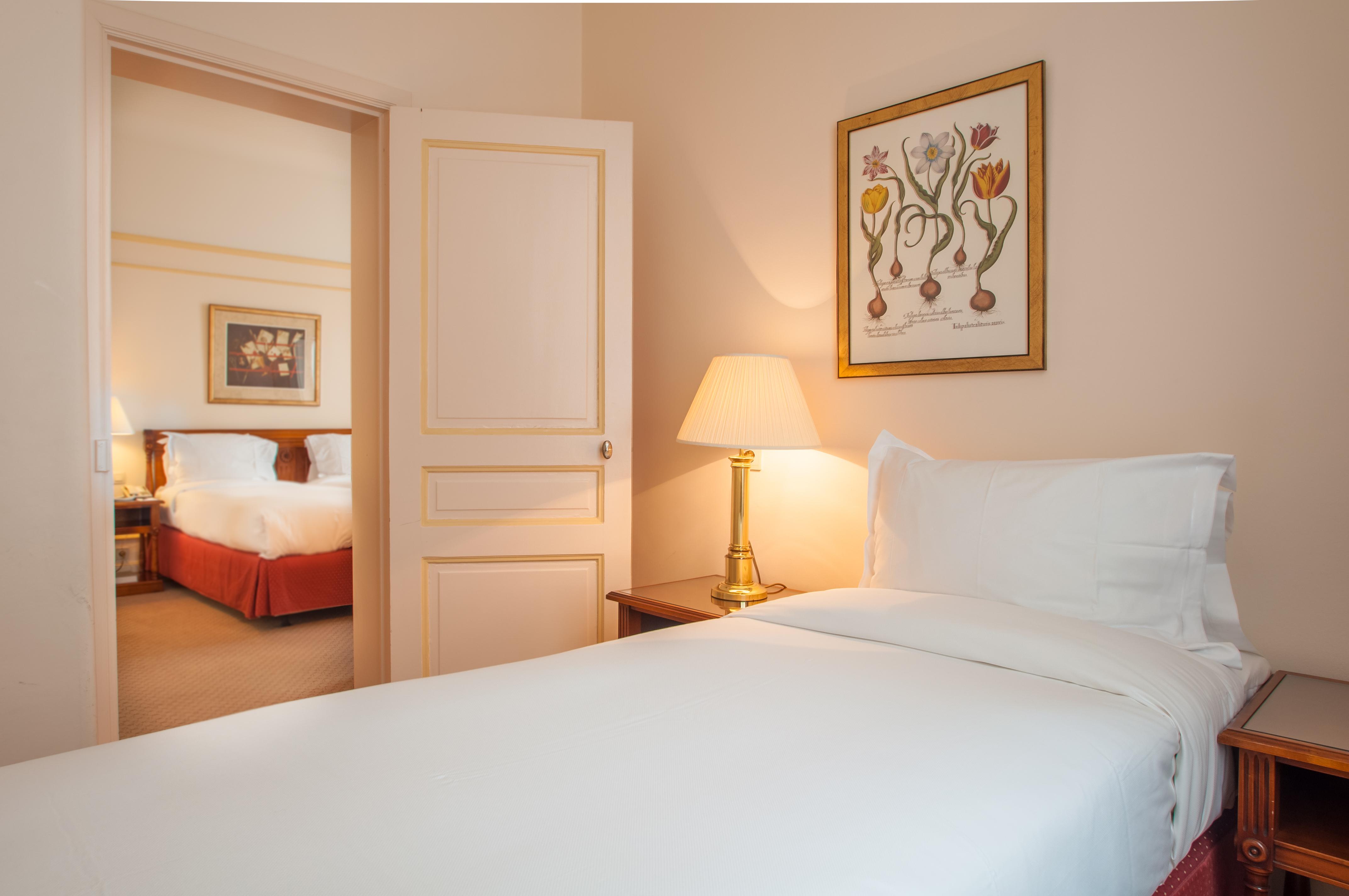 布鲁塞尔广场酒店  外观 照片 The photo shows a cozy hotel room with a single bed covered in white linens. There's a bedside table with a lamp providing soft light. On the wall, there is a framed piece of artwork featuring flowers. In the background, through an open door, a secon