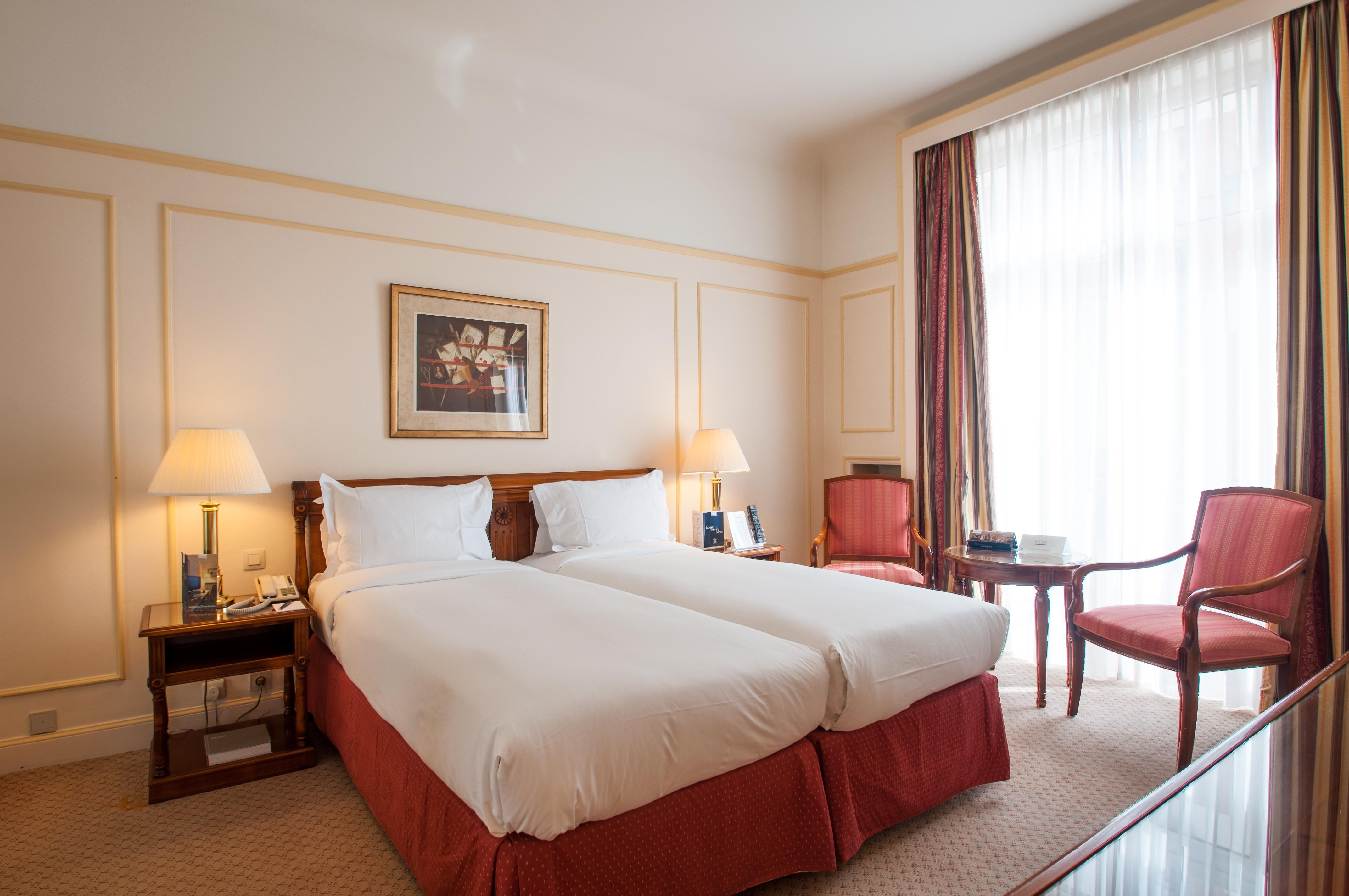 布鲁塞尔广场酒店  外观 照片 The photo shows a hotel room featuring two twin beds with white bedding and red accents. There are two bedside lamps on either side of the beds. In the background, there's a large window with sheer curtains allowing natural light to enter the room. T