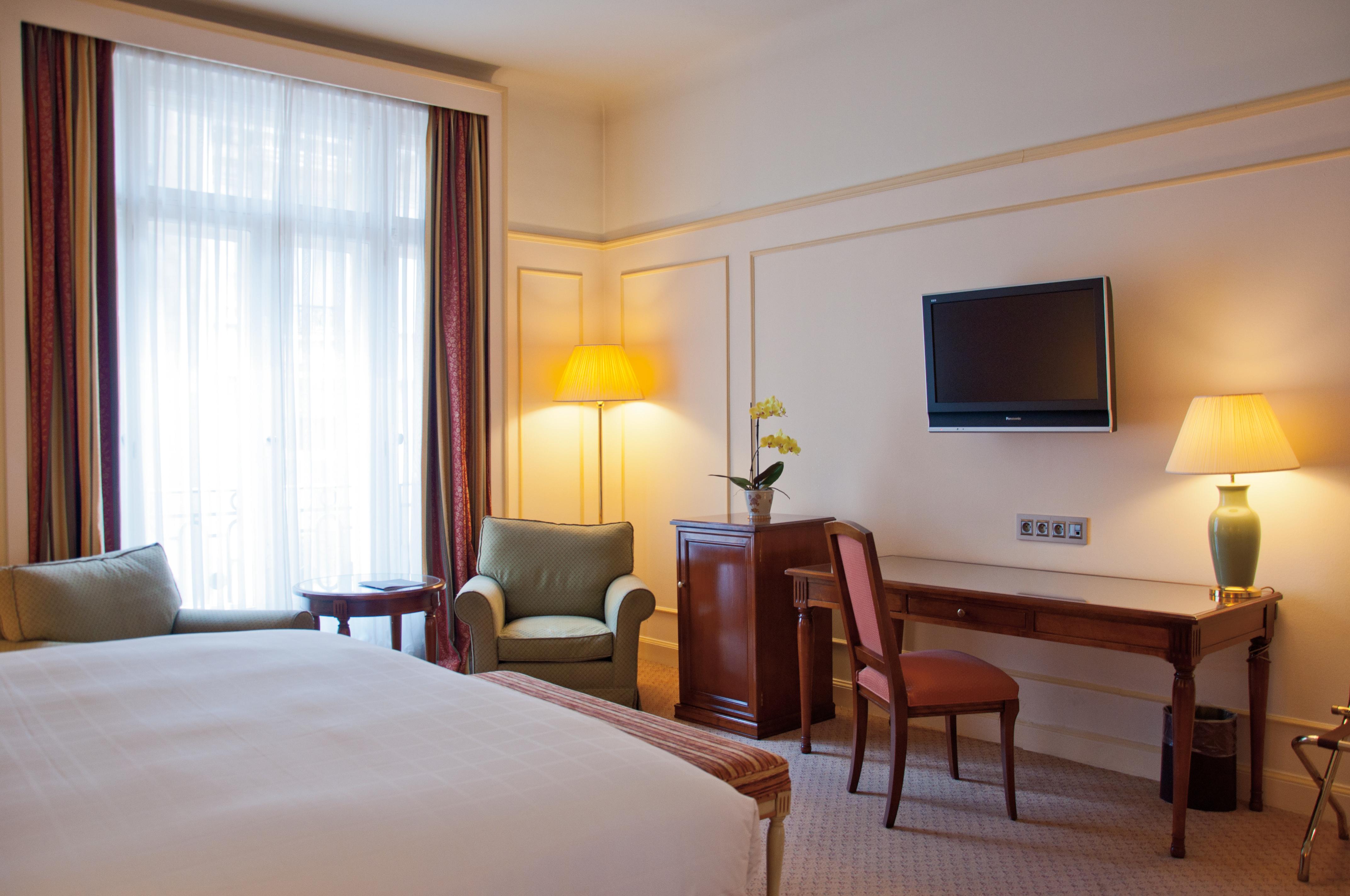 布鲁塞尔广场酒店  外观 照片 The photo depicts a hotel room featuring a large bed with white linens. On one side of the room, there is a small sitting area with a green armchair and a side table. A flat-screen TV is mounted on the wall opposite the bed. Next to the TV, there is 
