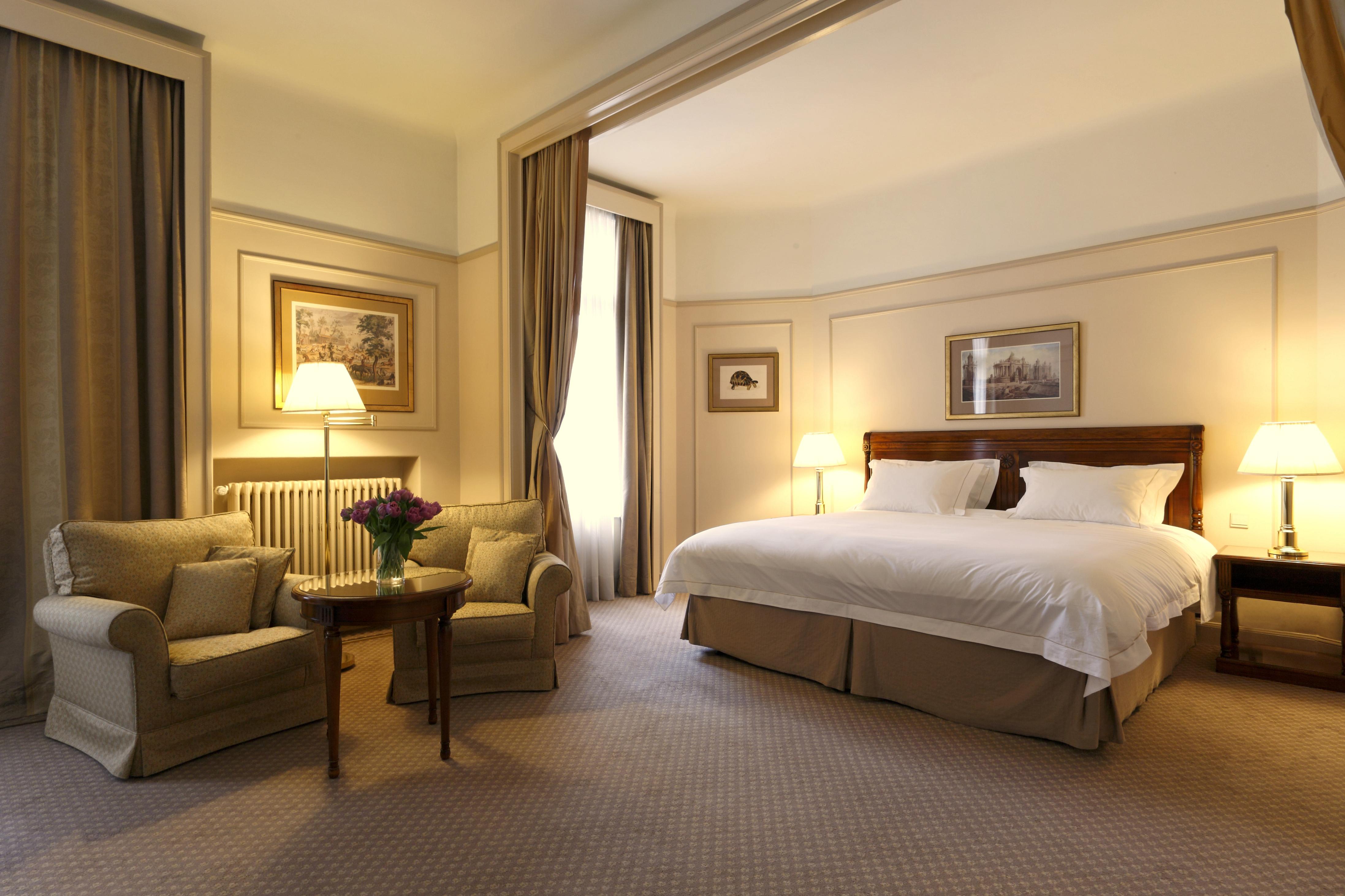 布鲁塞尔广场酒店  外观 照片 The photo shows a well-decorated hotel room featuring a large bed with white linens. There are two cozy armchairs positioned near a small round table. The room is illuminated by soft lighting from a floor lamp and wall sconces. Tall windows with floo