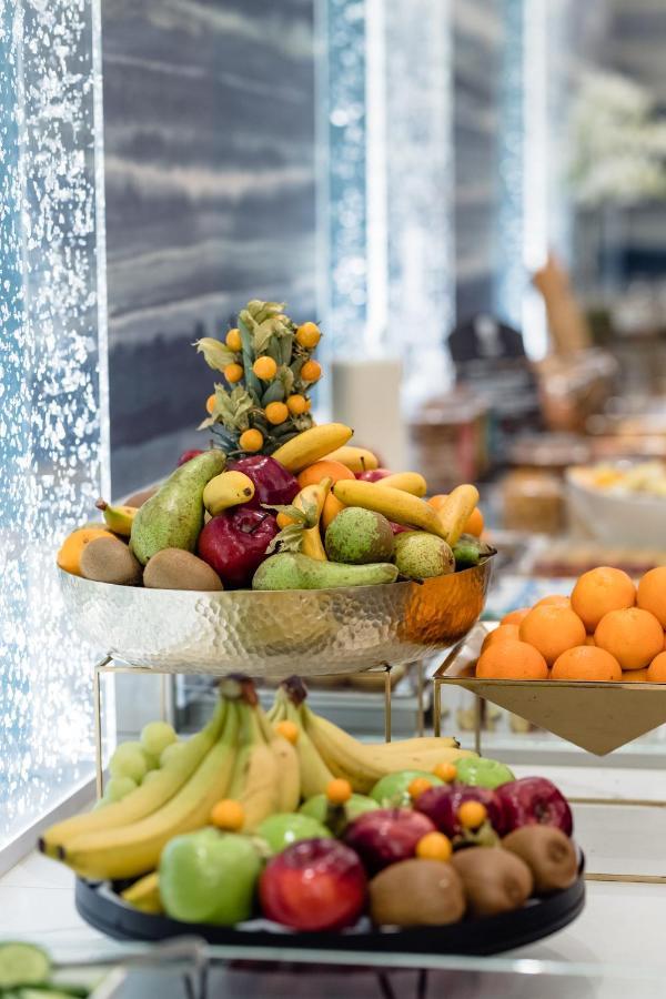 布鲁塞尔广场酒店  外观 照片 The photo shows a display of various fruits arranged in a bowl. The fruits include bananas, pears, apples, and some small citrus fruits, possibly kumquats, positioned attractively. The bowl is made of metal and has a shiny, textured surface. In the b