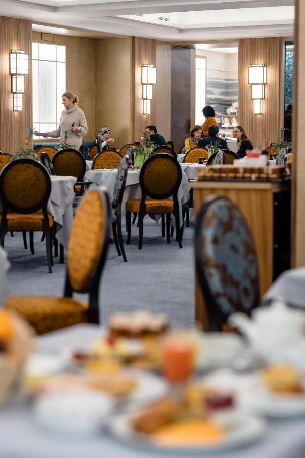 布鲁塞尔广场酒店  外观 照片 The photo depicts an elegant restaurant interior. There are several round tables covered with white tablecloths, each accompanied by ornate chairs featuring a gold and dark blue design. In the background, various groups of people are seated at differ
