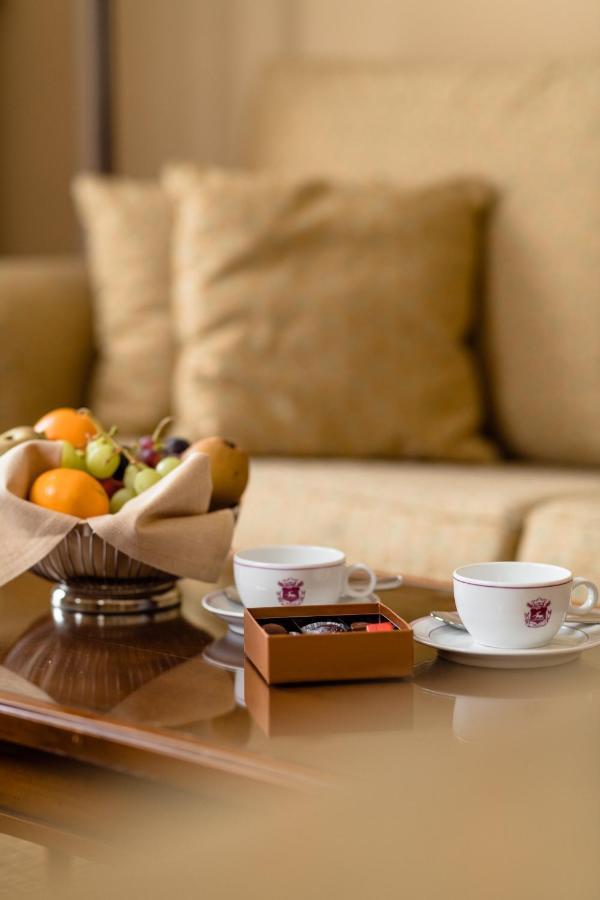 布鲁塞尔广场酒店  外观 照片 The photo shows a cozy setting with two small white cups placed on saucers, likely for coffee or tea. Next to the cups, there is a small box of chocolates. In the foreground, a silver bowl holds a variety of fruits, including oranges, grapes, and oth