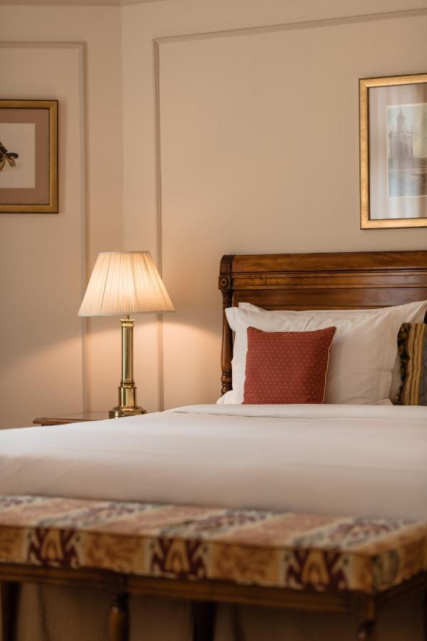 布鲁塞尔广场酒店  外观 照片 The photo shows a cozy bedroom setting. There is a wooden bed with white bedding and a decorative red pillow. Next to the bed, there is a bedside lamp with a cream-colored lampshade. The walls are adorned with subtle decorations, likely framed artwor