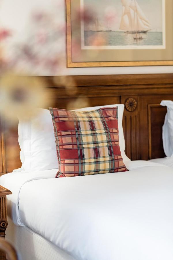 布鲁塞尔广场酒店  外观 照片 The photo shows a decorative plaid pillow placed on a bed. The pillow features a mix of red, yellow, and dark plaid patterns. The bedding is white, and there is a wooden headboard in the background, which has a decorative design. The overall ambiance
