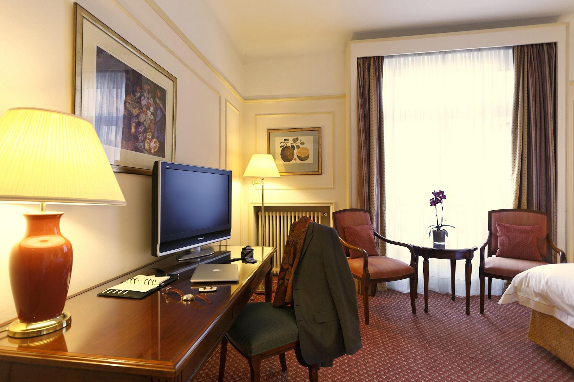布鲁塞尔广场酒店  外观 照片 The photo shows a well-furnished hotel room. Key features include:

- A desk with a laptop and some stationery items.
- A flat-screen television mounted on the wall.
- A bedside lamp providing warm light.
- Two comfortable chairs around a small circu