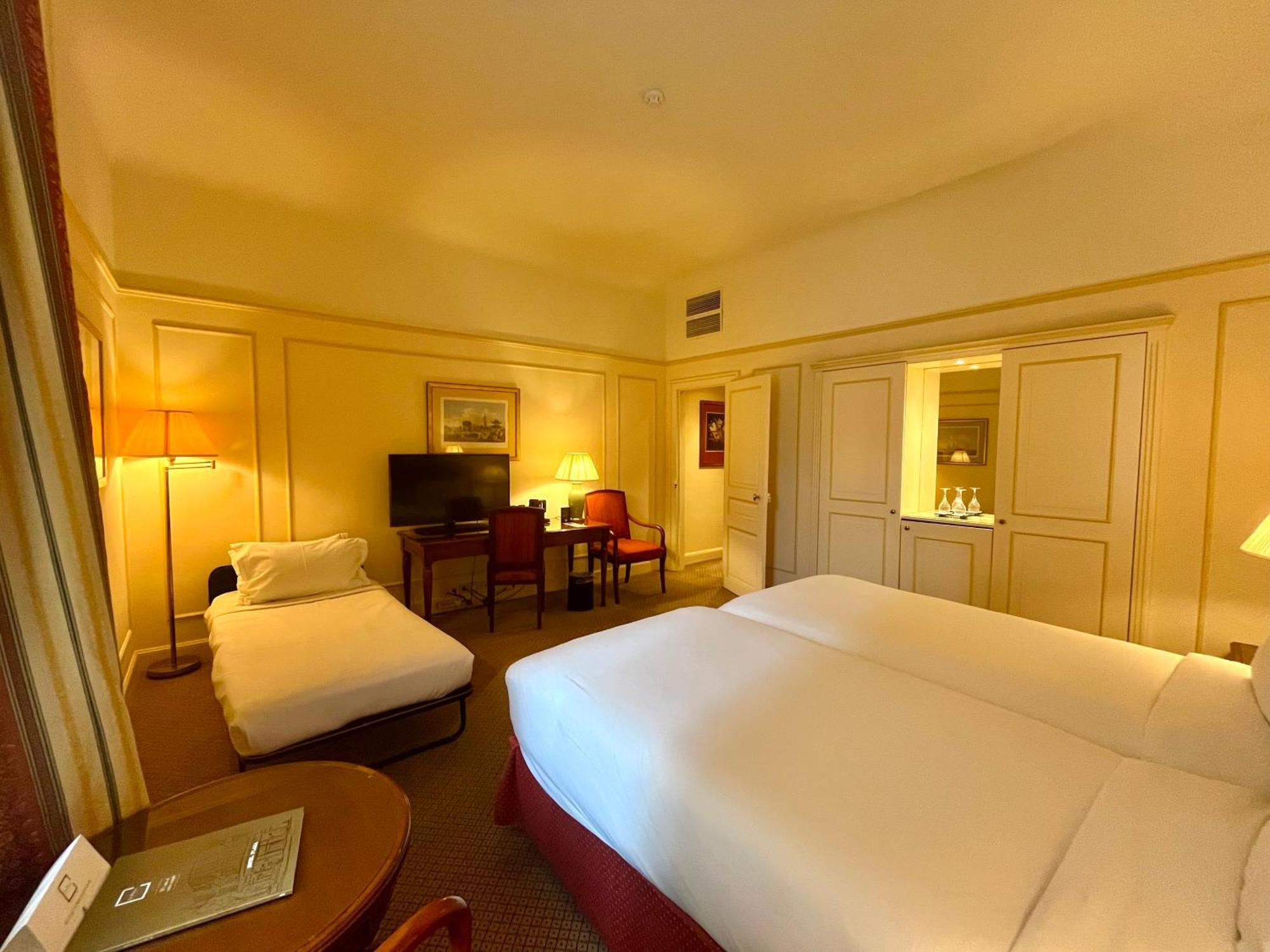 布鲁塞尔广场酒店  客房 照片 The photo shows an interior of a hotel room with a warm, cozy ambiance. It features two single beds made with white linens positioned side by side. There is a small, rolled-up cot or mattress in the corner. Near the beds, there is a bedside table and