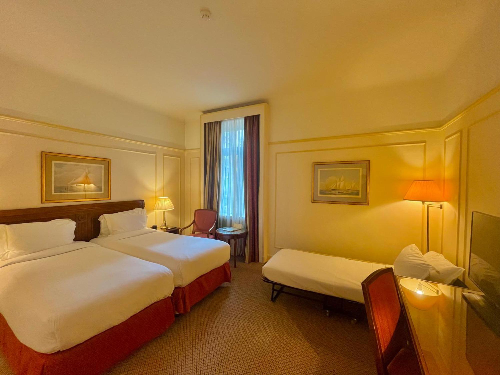 布鲁塞尔广场酒店  客房 照片 The photo shows a hotel room featuring two single beds with white linens and red accents. There is a small extra bed on the floor, along with a wooden chair. A desk and a lamp are present near the window, which has sheer curtains allowing natural lig
