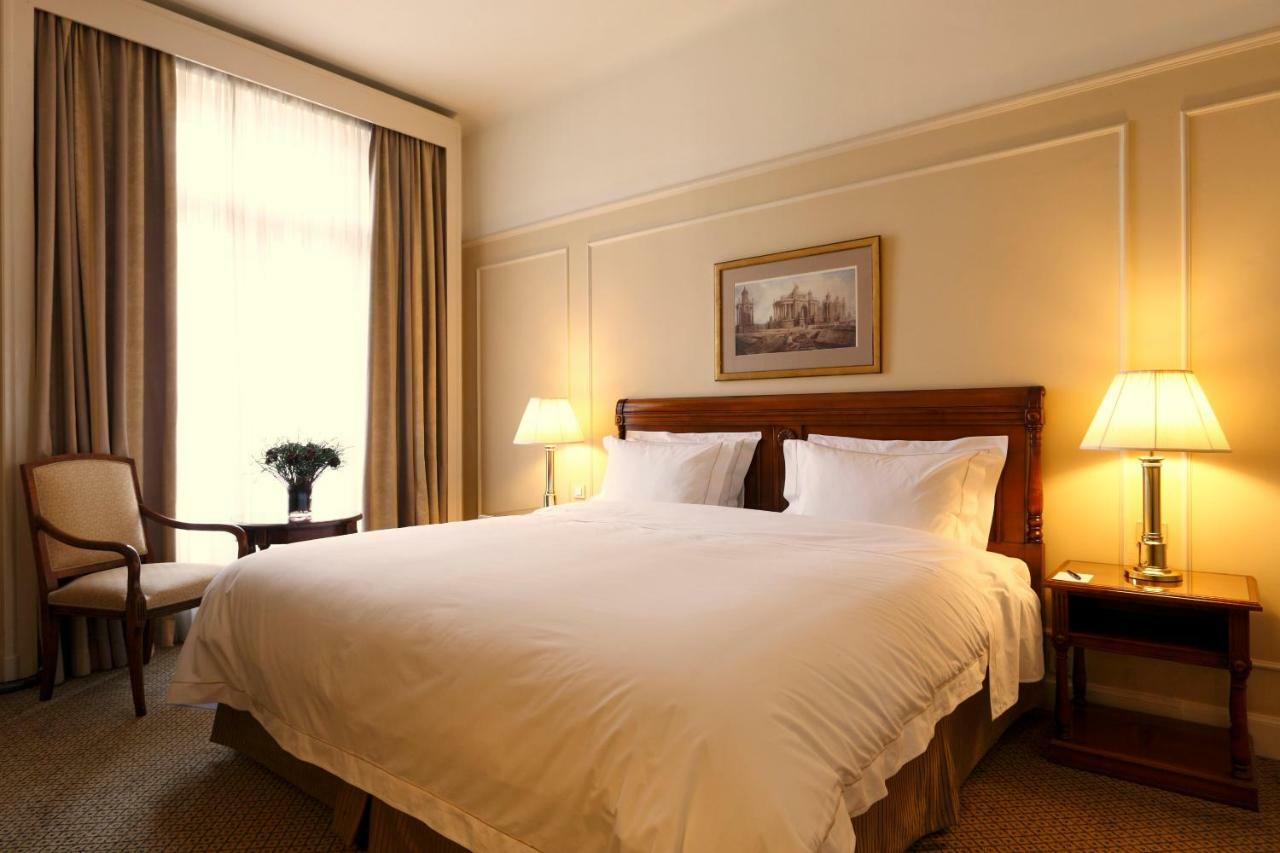 布鲁塞尔广场酒店  外观 照片 The photo shows a well-decorated hotel room featuring a large bed with white bedding. There are two bedside lamps providing warm illumination. A small wooden table sits beside a chair, and there's a potted plant on the table. Natural light enters thr